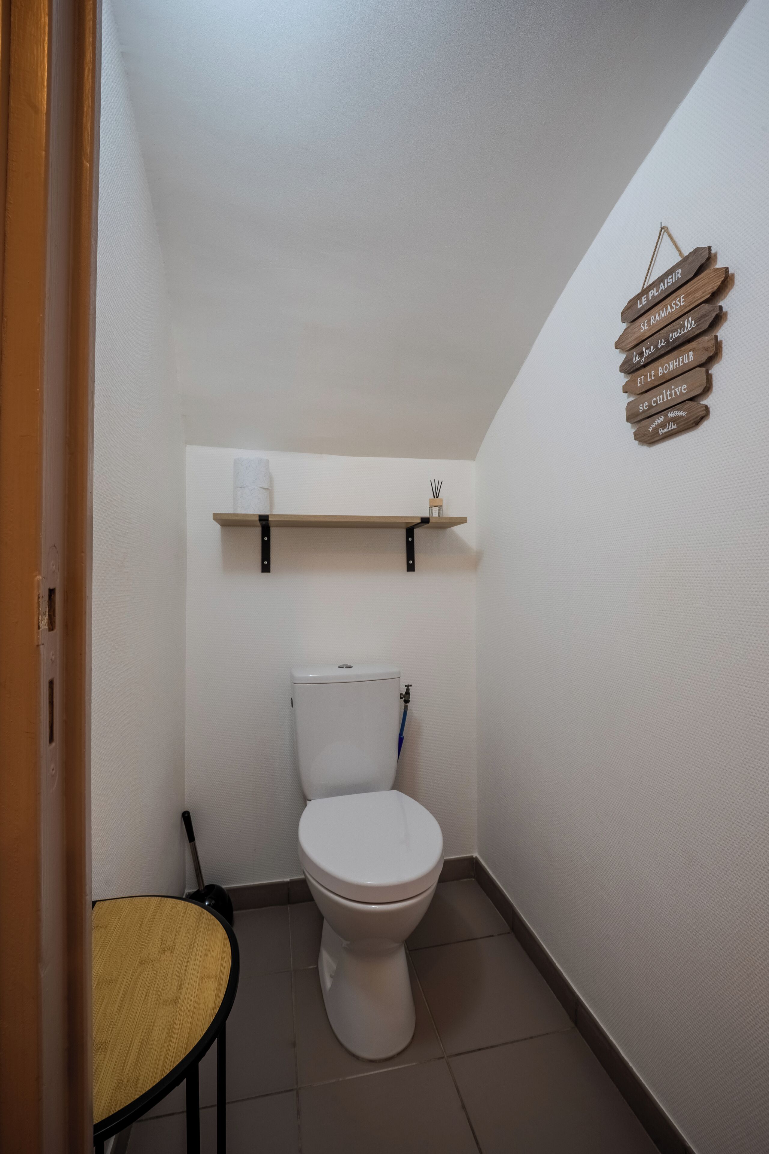 A small, clean bathroom with a toilet, a shelf above the toilet, and a small table.