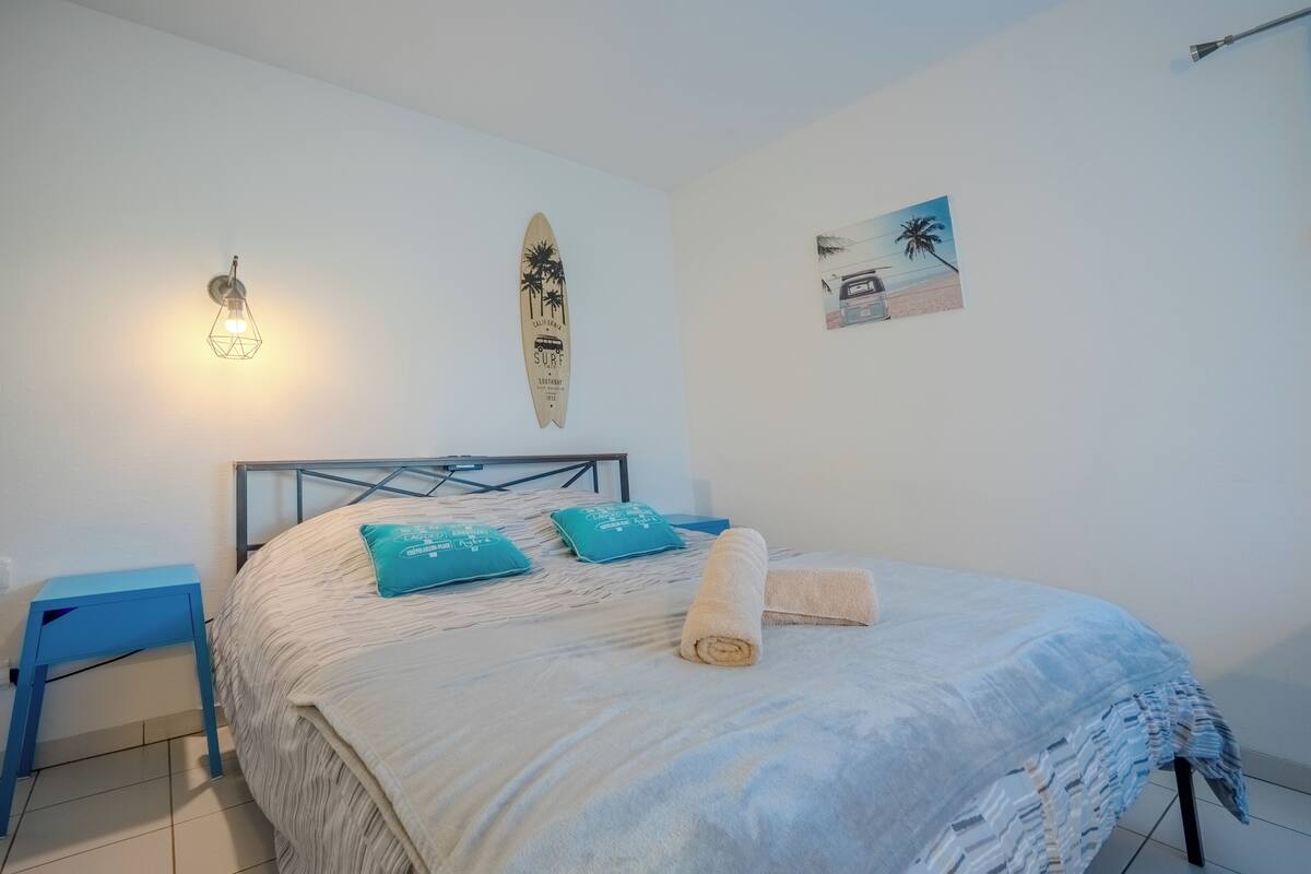 A neatly made bed with a white and blue striped blanket, two teal pillows, and a rolled towel. A wall-mounted light fixture and a surfboard decoration are visible.