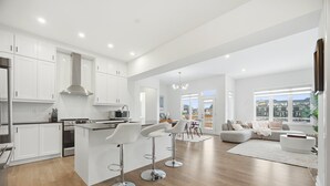 A modern kitchen with white cabinets and a central island, a dining area with a white table and chairs, and a living room with a grey sofa and a large window.