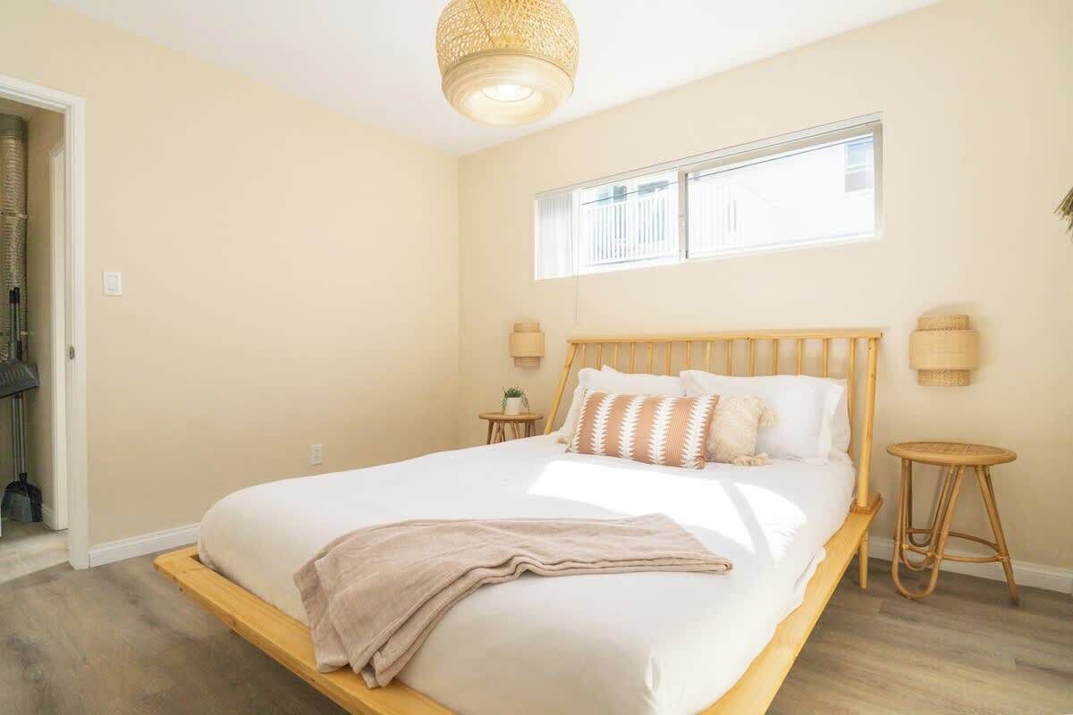 Quarto com cama grande, cabeceira de madeira, duas mesas de cabeceira, uma janela e um lustre.