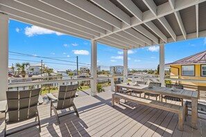 Um deck coberto ao ar livre com móveis de madeira, vista para o porto de iates, e um prédio amarelo ao fundo.