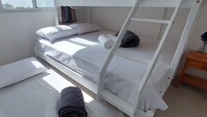 Habitación con literas, ropa de cama blanca, una mesita de noche de madera y ventana con vistas a edificios.