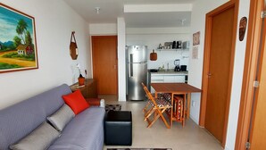 Una sala de estar con sofá, una mesa de comedor con sillas, una zona de cocina con refrigerador y una puerta que da a otra habitación.
