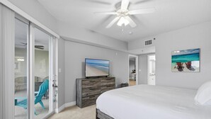 A bedroom with a large bed, a ceiling fan, a TV on a wooden dresser, and a sliding glass door leading to a balcony. - Oceanwalk 12-302 (New Smyrna Beach)
