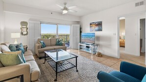 Ein großzügiges Wohnzimmer mit einer Couch, einem Couchtisch und einem Fernseher. Ein Balkon bietet Blick auf den Ozean.