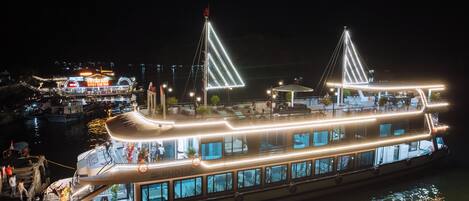 A large, illuminated boat with multiple decks, featuring a prominent bridge structure and a dining area with outdoor seating.