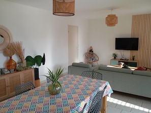 Una sala de estar con un sofá, una mesa de comedor con mantel estampado, un televisor en la pared y una lámpara en el techo.