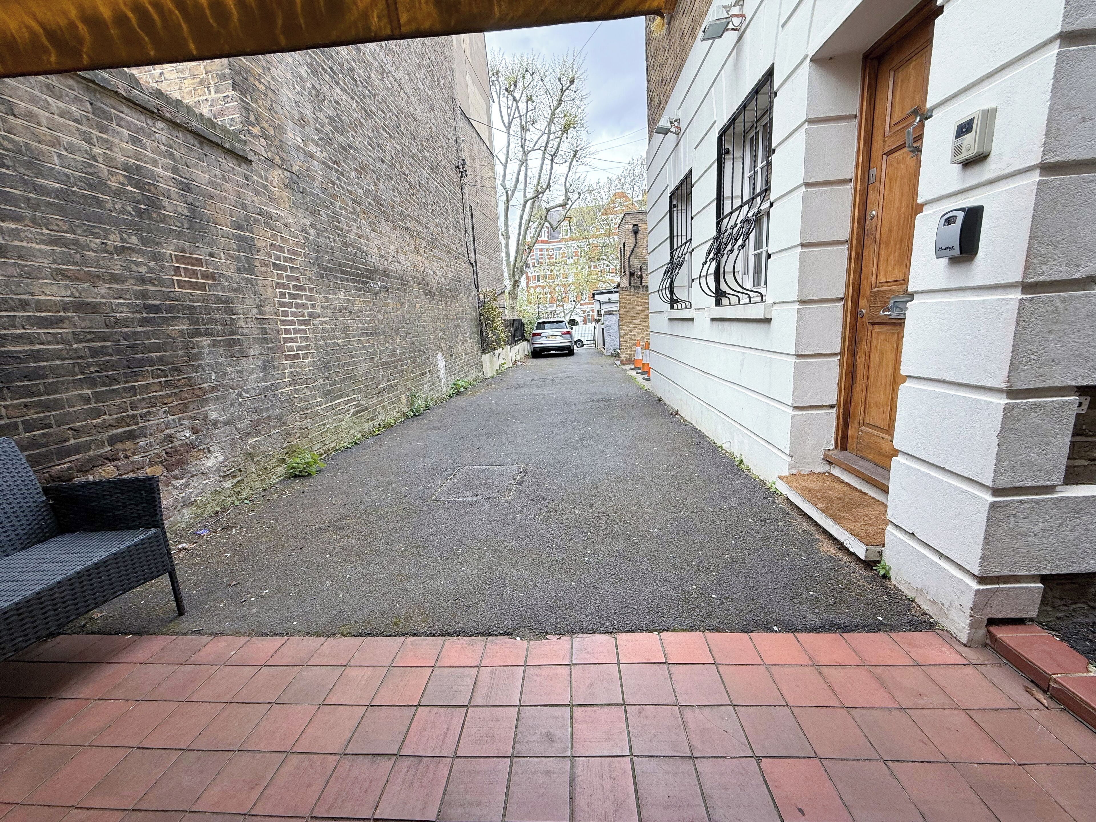 A narrow alleyway with a brick wall on one side and a white building with a wooden door on the other. There is a bench on the left and a car parked in the distance.
