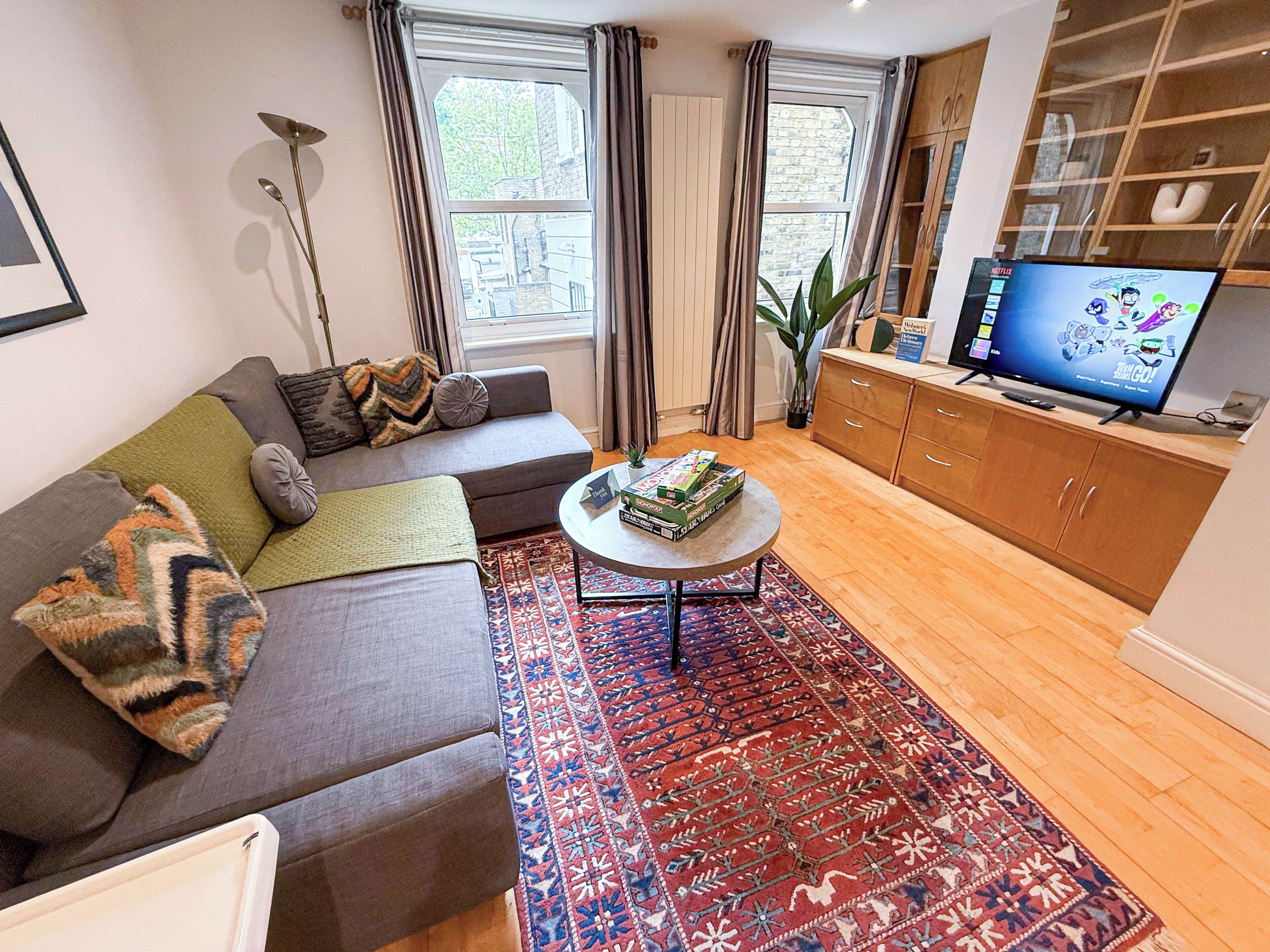 Un salon avec un canapé gris, une table basse sur laquelle sont posés des jeux de société, un téléviseur sur un meuble en bois et un tapis à motifs.
