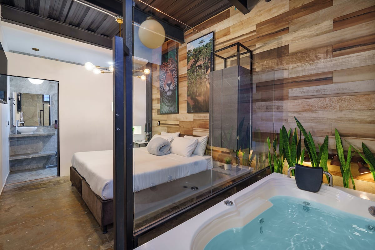 A modern bedroom with a glass-enclosed bed, a bathroom with a large mirror, and a hot tub.