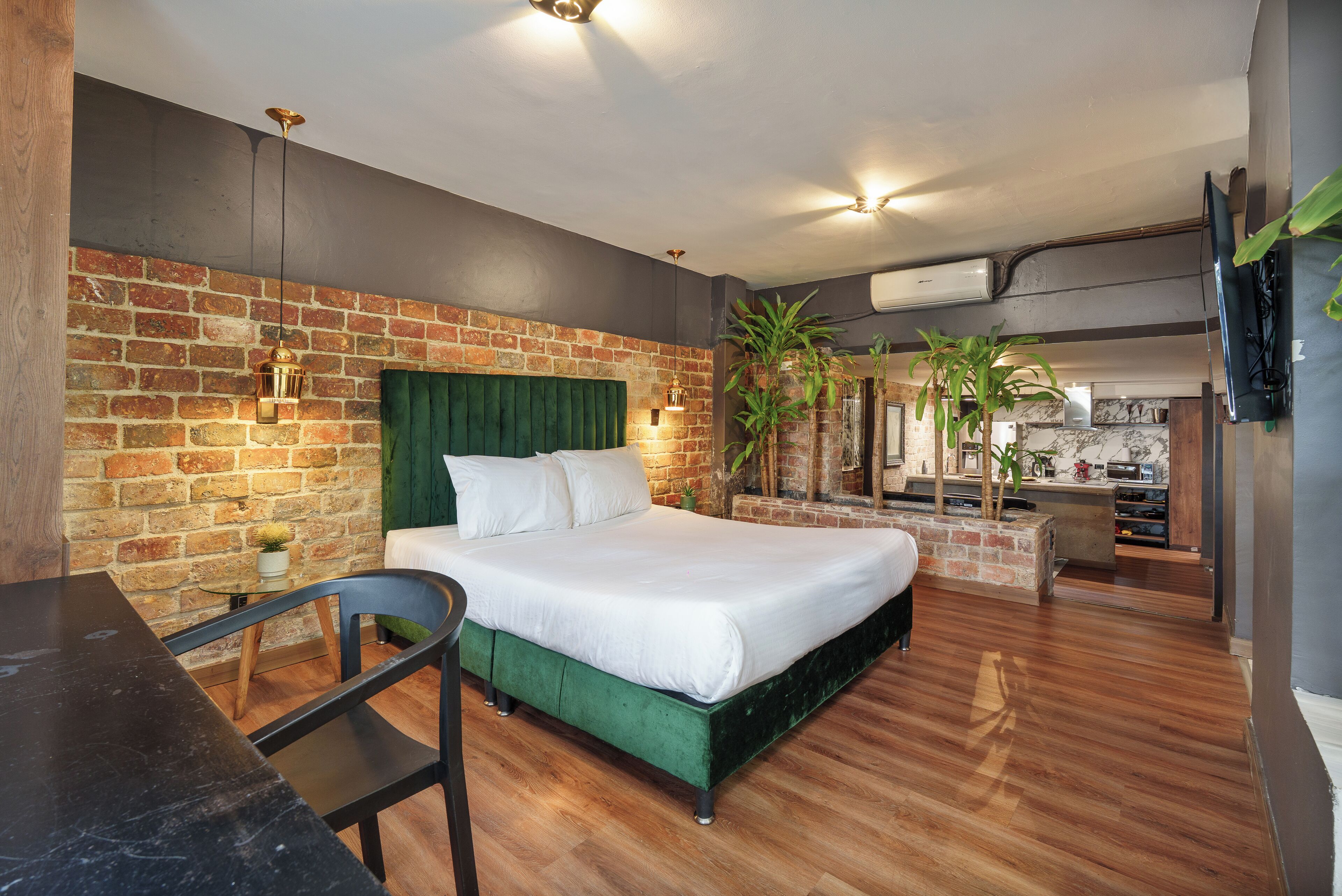 A modern hotel room with a brick wall, a green upholstered bed, a wooden desk, and a small plant.