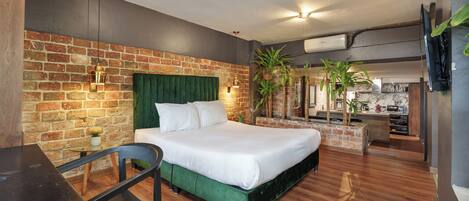 A modern hotel room with a brick wall, a green upholstered bed, a wooden desk, and a small plant.