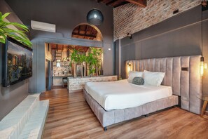 A modern bedroom with a large bed, wooden flooring, a brick wall, and a cityscape painting. - Lleras Vibex Party (Medellín)