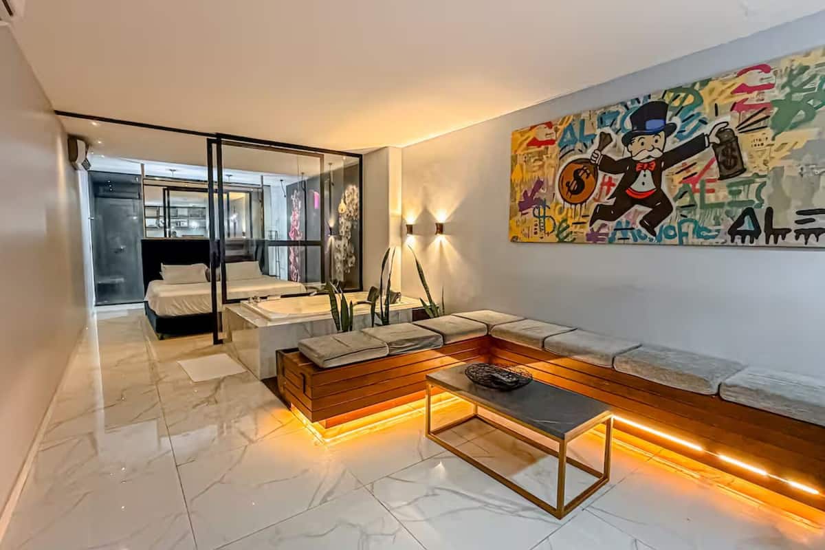 A modern bedroom with a glass door leading to a bathroom, a wooden bench with cushions, and a colorful abstract painting on the wall.