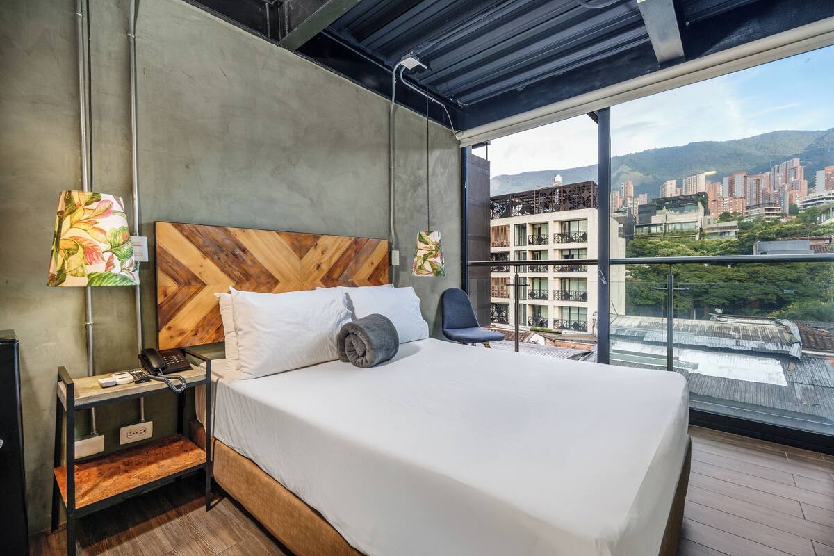 A modern hotel room with a large bed, a wooden headboard, a nightstand with a phone, and a view of the city through large windows.