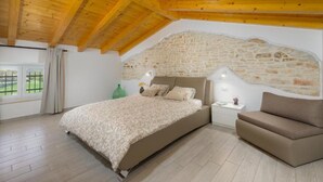 Ein Schlafzimmer mit schrägem Dach, einer Steinwand, einem Bett, einem Nachttisch und einem Sessel.