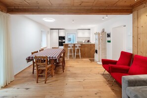 Zona pranzo con tavolo e sedie in legno, una poltrona rossa e una cucina sullo sfondo.
