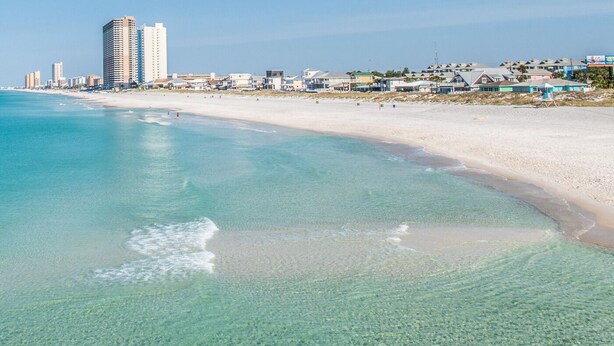 Ein Strand mit klarem türkisblauem Wasser, einem sandigen Ufer und Gebäuden im Hintergrund.