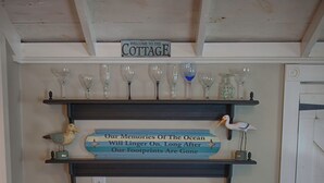 Une pièce chaleureuse avec une table ronde en bois et deux chaises, une étagère avec des verres à vin et un oiseau décoratif, et une pancarte au-dessus indiquant : « BIENVENUE AU COTTAGE ».