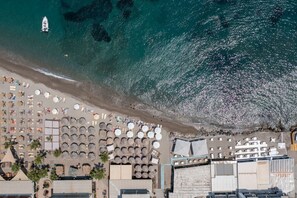 An aerial view of a beach with rows of umbrellas and sun loungers, a boat on the water, and buildings adjacent to the shore. - Aram`s Luxury Port House (Kos)