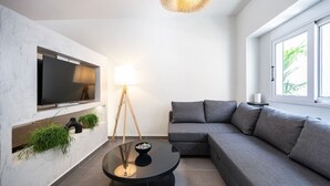 Ein modernes Wohnzimmer mit großem Flachbildfernseher an einer weißen Wand, einem grauen Sofa, einem runden schwarzen Couchtisch und einer Stehlampe.
