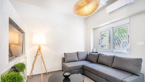 Ein modernes Wohnzimmer mit einem grauen Ecksofa, einem runden schwarzen Couchtisch, einer Stehlampe und einem Fenster mit Blick auf Grünflächen.