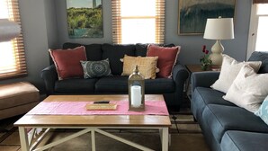 Ein Wohnzimmer mit einem dunkelblauen Sofa, einem Holztisch und einem rosa Teppich. An der Wand hängen zwei Gemälde, auf einem Beistelltisch steht eine Lampe und es gibt ein Fenster mit Jalousien.