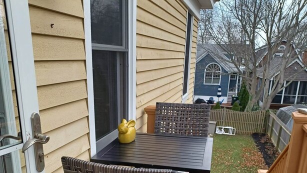 Eine überdachte Terrasse mit Tisch und Stühlen, ein gelbes Dekorationsobjekt und Blick auf einen Garten mit Zaun und Bäumen.