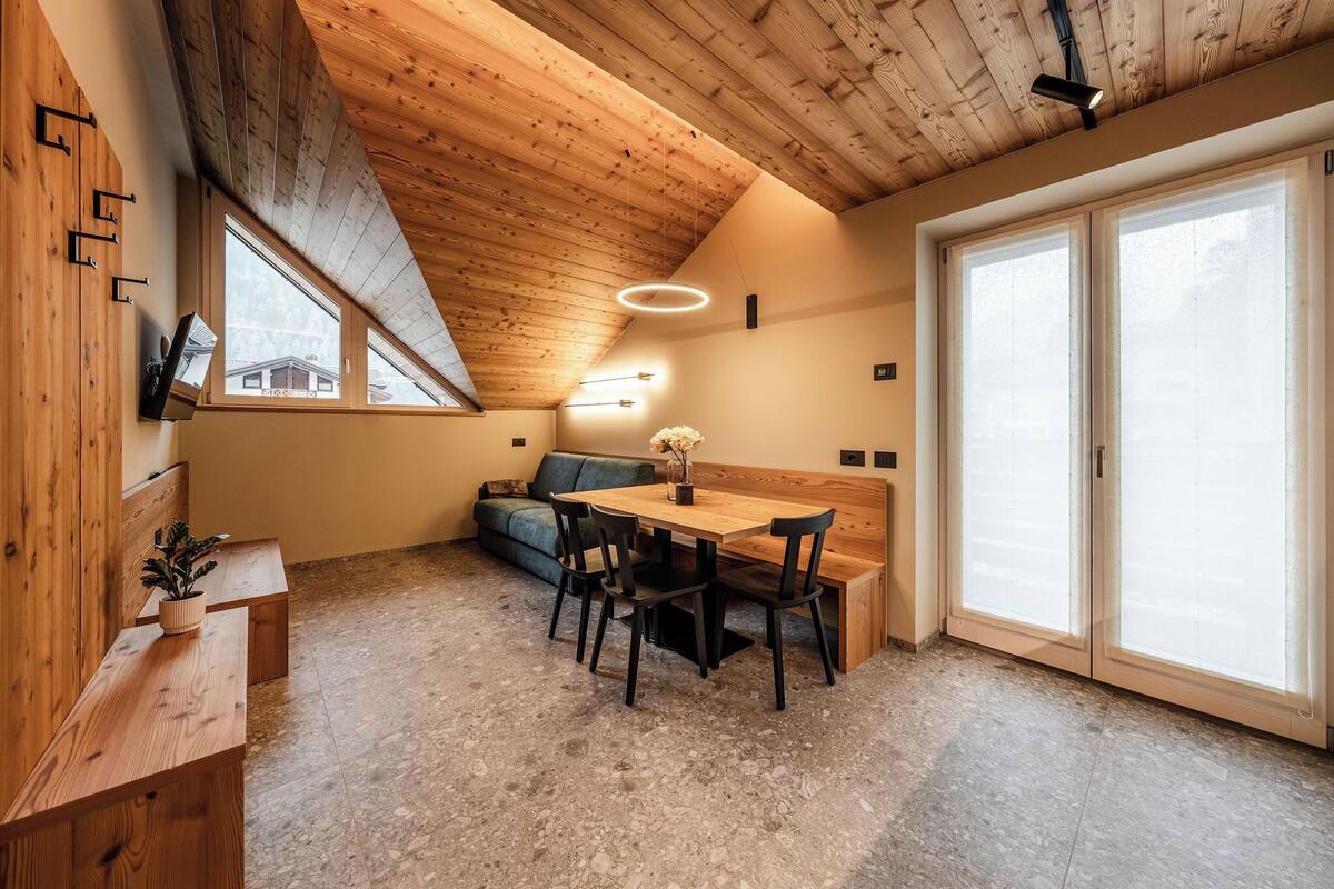 A cozy room with a sloped ceiling, a wooden dining table with chairs, a bench, and a sofa. There are windows and a door with frosted glass.