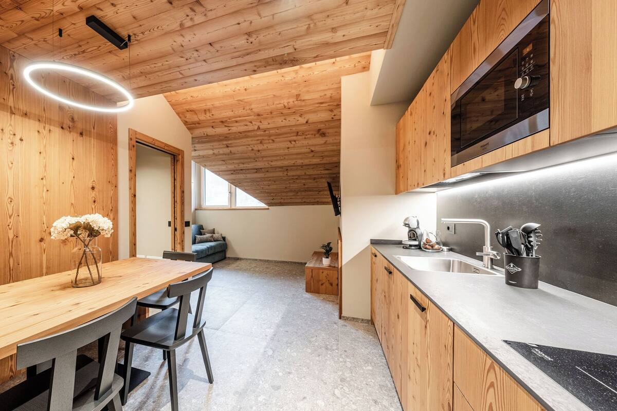 A modern kitchen with wooden cabinetry, a stainless steel refrigerator, and a built-in oven. There is a dining area with a wooden table and chairs, and a small plant on a side table.