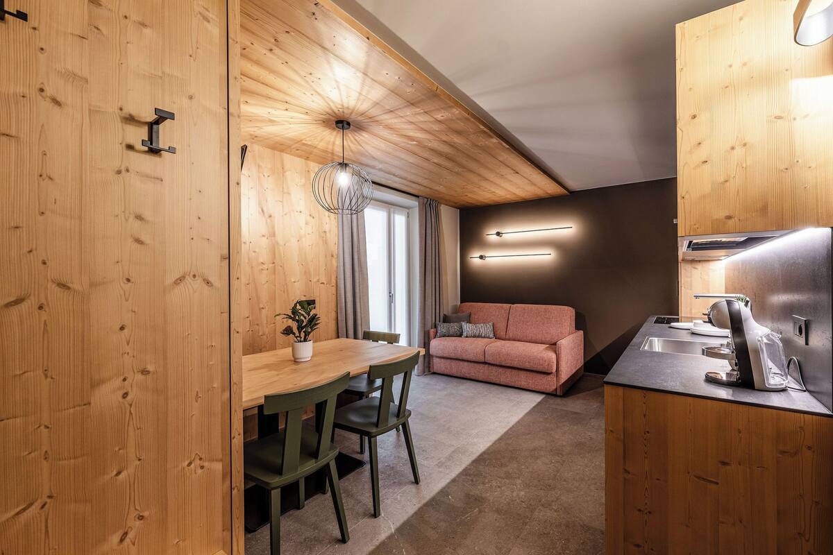 A compact kitchen with wooden cabinetry, a dining area with a table and chairs, a sofa, and a modern pendant light.