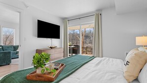 Ein modernes Schlafzimmer mit einem großen Bett, einem an der Wand montierten Fernseher, einem Schreibtisch und Blick ins Freie durch eine Glasschiebetür.