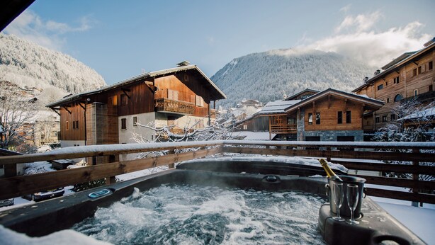 Una cabaña de madera con bañera de hidromasaje y balcón, rodeada de edificios nevados y montañas.