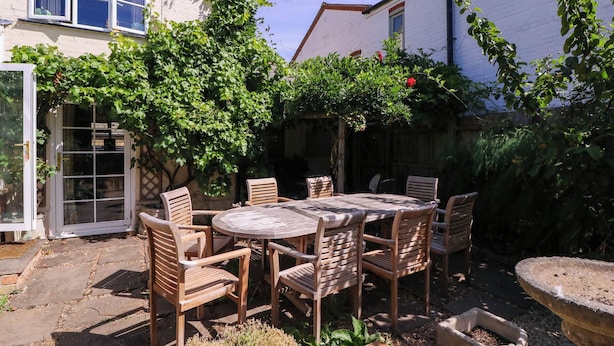 A patio with a round table and six wooden chairs, surrounded by greenery and a white building. - Vine Cottage (Tewkesbury)