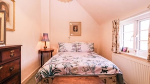 A bedroom with a bed, a dresser, a bedside table, a lamp, a picture frame, and a window with curtains. - Vine Cottage (Tewkesbury)