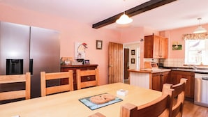 A kitchen with wooden cabinets, a stainless steel refrigerator, a wooden dining table with chairs, and a pendant light. - Vine Cottage (Tewkesbury)