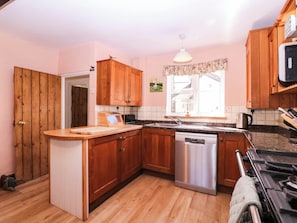 A kitchen with wooden cabinets, a built-in dishwasher, a countertop with a toaster, and a window with a view. - Vine Cottage (Tewkesbury)
