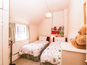 A bedroom with two beds, a window with curtains, a radiator, a bookshelf with books and stuffed animals, and a teddy bear on a dresser. - Vine Cottage (Tewkesbury)