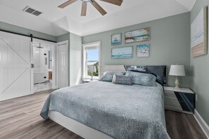 A bedroom with a bed, a nightstand, a ceiling fan, artwork on the wall, and a window with a view. - Outer Banks Salvo Family Retreat: Private Pool | Private Hot Tub | Dog Friendly (Salvo)