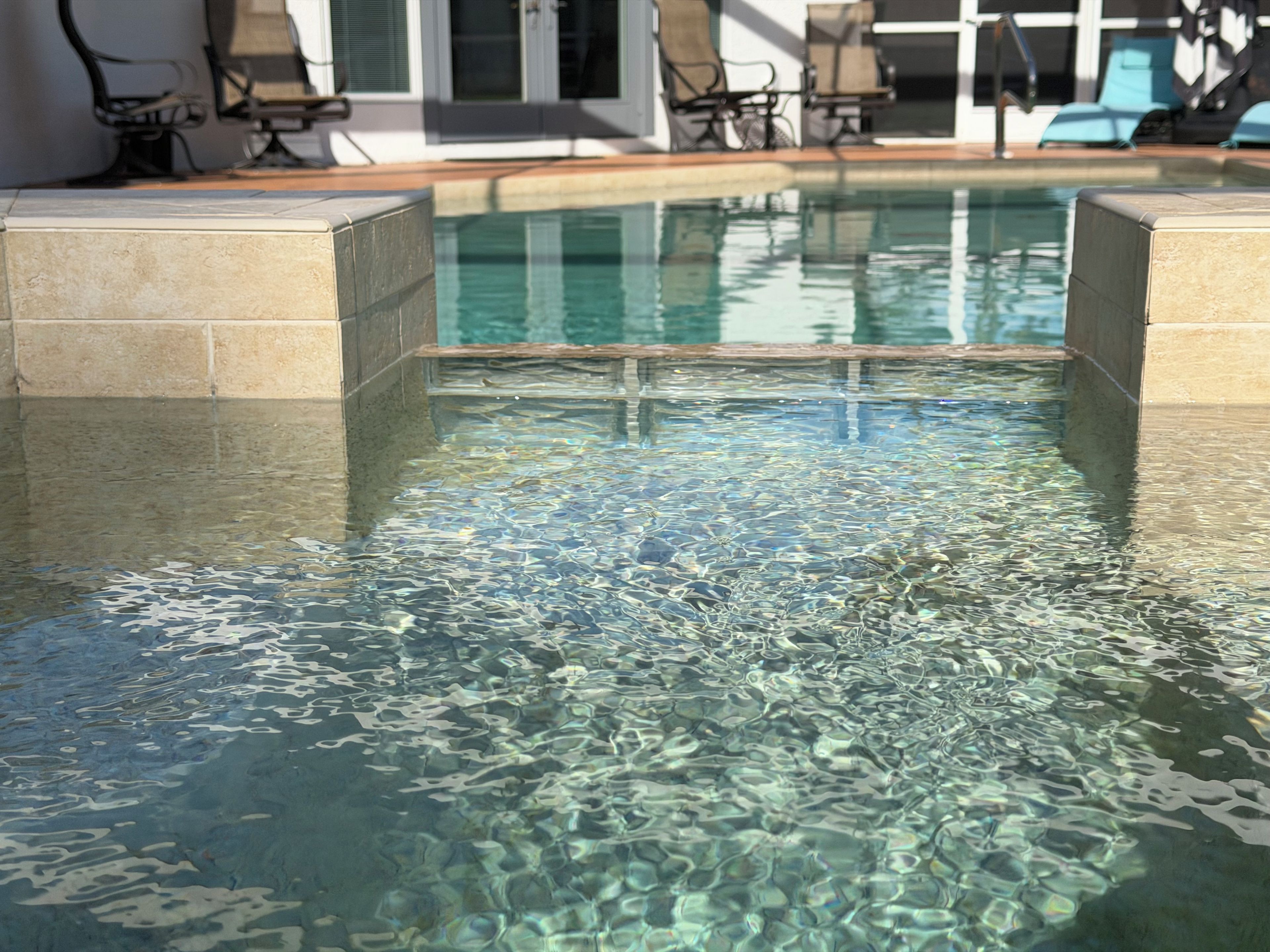 A clear swimming pool with a tiled edge, surrounded by lounge chairs and a building in the background.