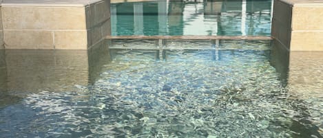 A clear swimming pool with a tiled edge, surrounded by lounge chairs and a building in the background.