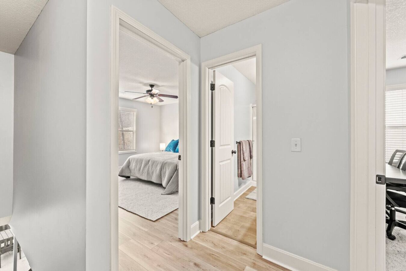 Ein Flur, durch eine offene Tür ist ein Schlafzimmer sichtbar, mit einem Bett, einem Deckenventilator und einem Fenster mit Jalousien.