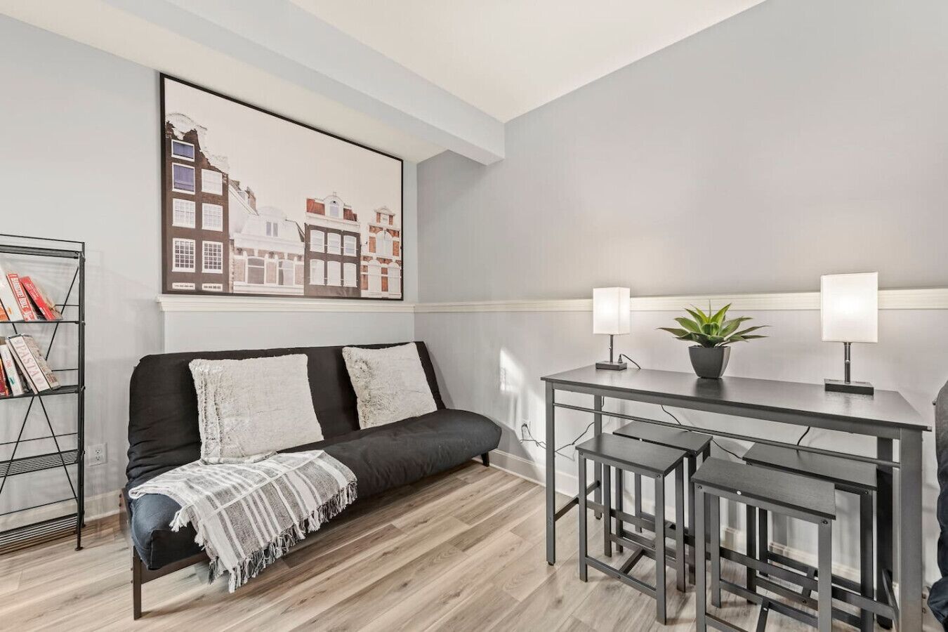 Un salon moderne avec un canapé noir, une table à manger avec deux chaises, une bibliothèque et un tableau représentant un paysage urbain accroché au mur.