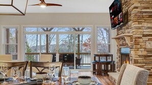 A dining area with a wooden table set for a meal, a stone fireplace, and a large window overlooking a deck. - Lakefront • HotTub • Luxe Baths • Fire Pit Retreat (Mineral)