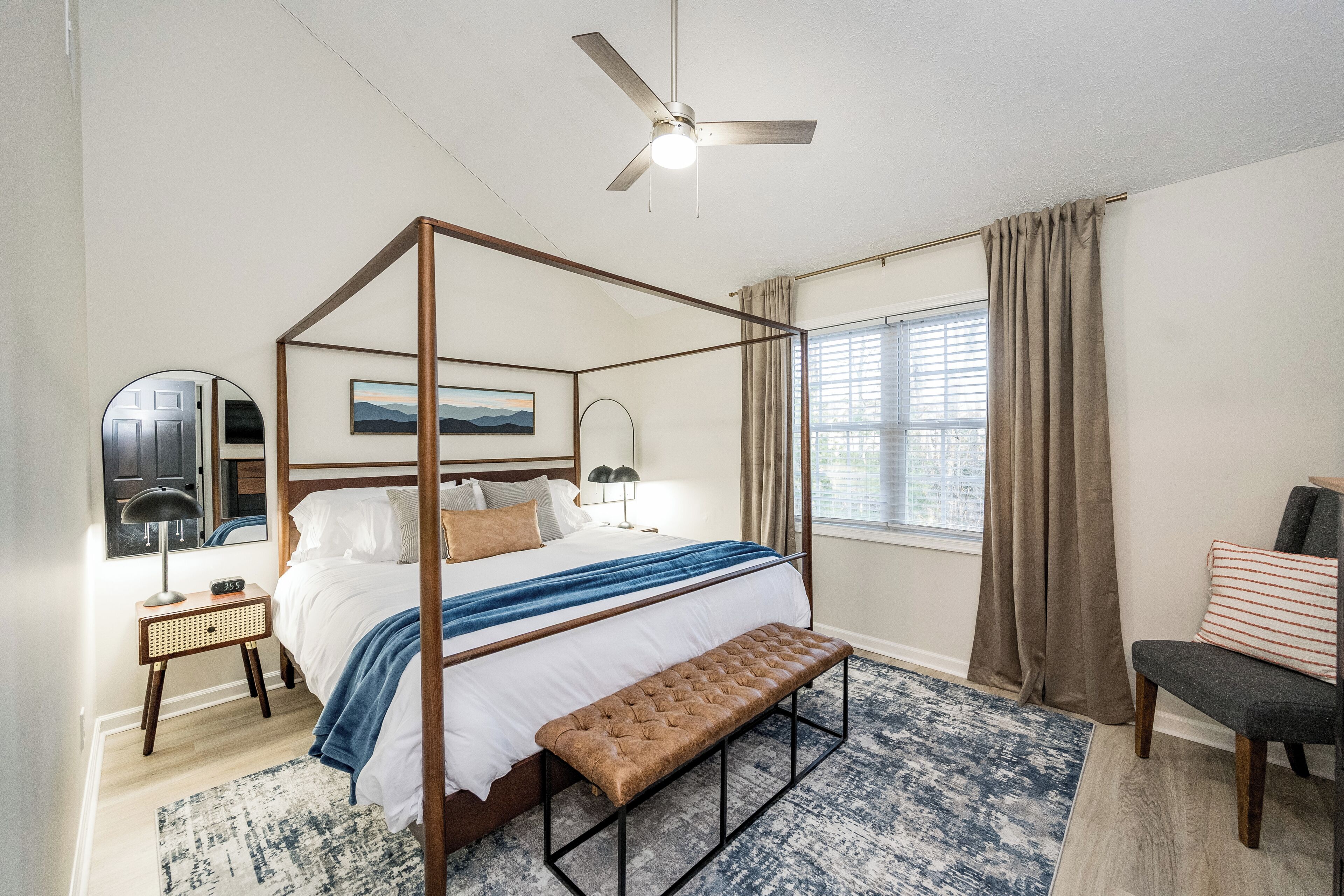 A four-poster bed with a bench, a ceiling fan, a bedside table with a lamp, a window with curtains, and a chair.