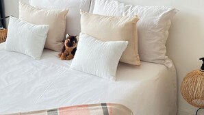 Een net opgemaakt bed met witte beddengoed en meerdere kussens, een kleine hond op het bed, en een geweven nachtkastje.
