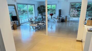 Un vaste espace de vie comprenant une table et des chaises en verre pour la salle à manger, une grande plante en pot et une vue sur un balcon doté d’une balustrade.