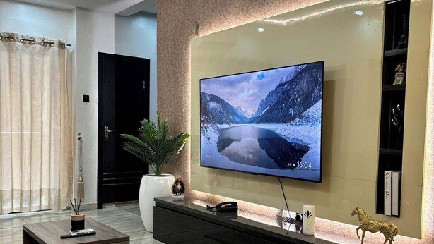 A modern living room with a large flat-screen TV mounted on the wall, a dark wooden coffee table, a potted plant, and a decorative horse figurine. - Icempire Signature (Lagos)