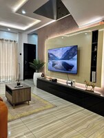 Uma sala de estar moderna com uma grande televisão de tela plana na parede, uma mesa de centro de madeira escura, um vaso de plantas e uma pequena estatueta de cavalo decorativa.