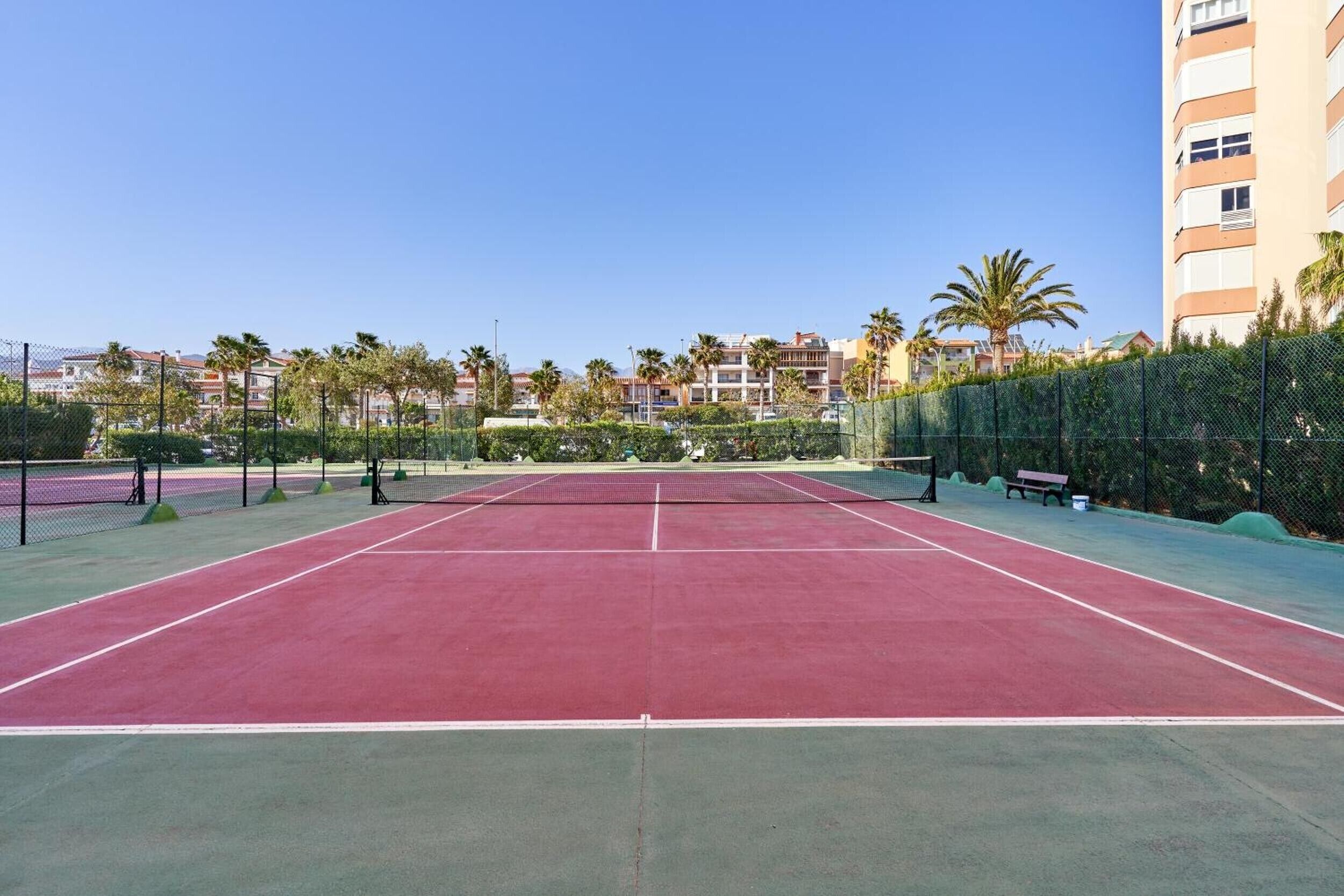 A tennis court with a net, surrounded by a fence, and buildings in the background.